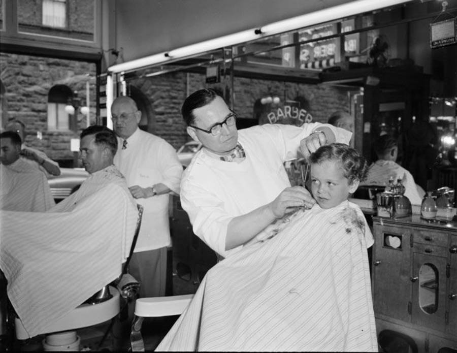 A barber cuts a young boy’s hair while another barber works on an adult in an old-fashioned barbershop. The shop’s large windows and vintage decor are visible in the background.