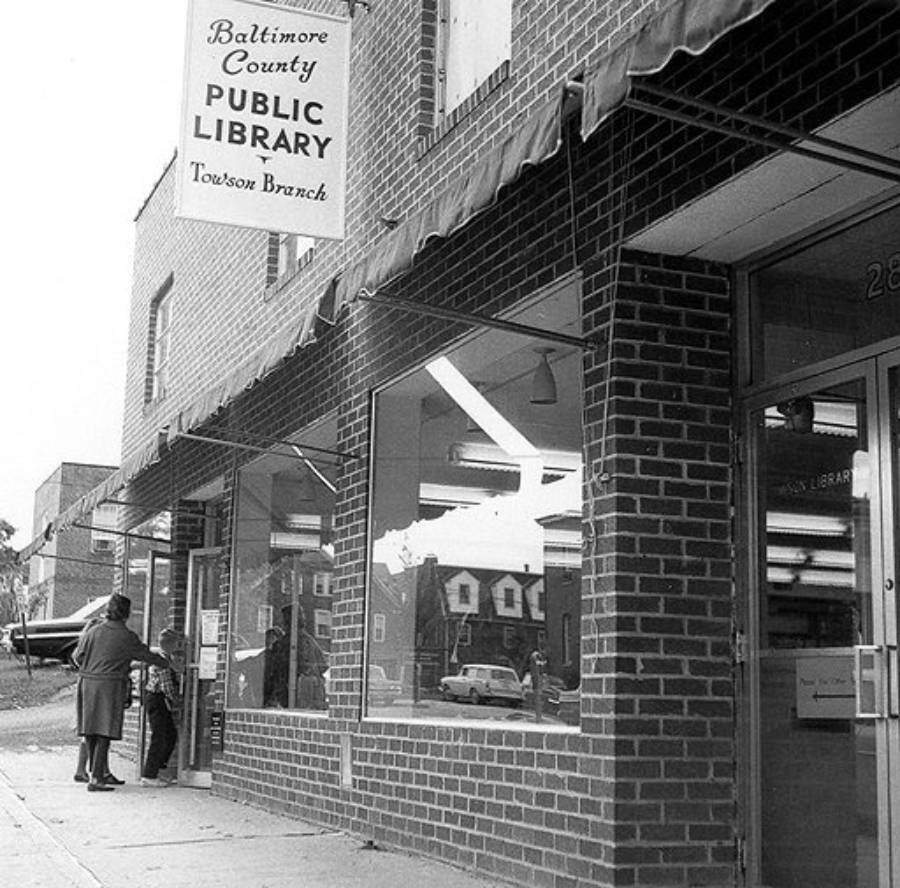 A person stands outside the Baltimore County Public Library, Towson Branch. The brick building has large windows and a sign above the entrance. The street and nearby buildings are visible.