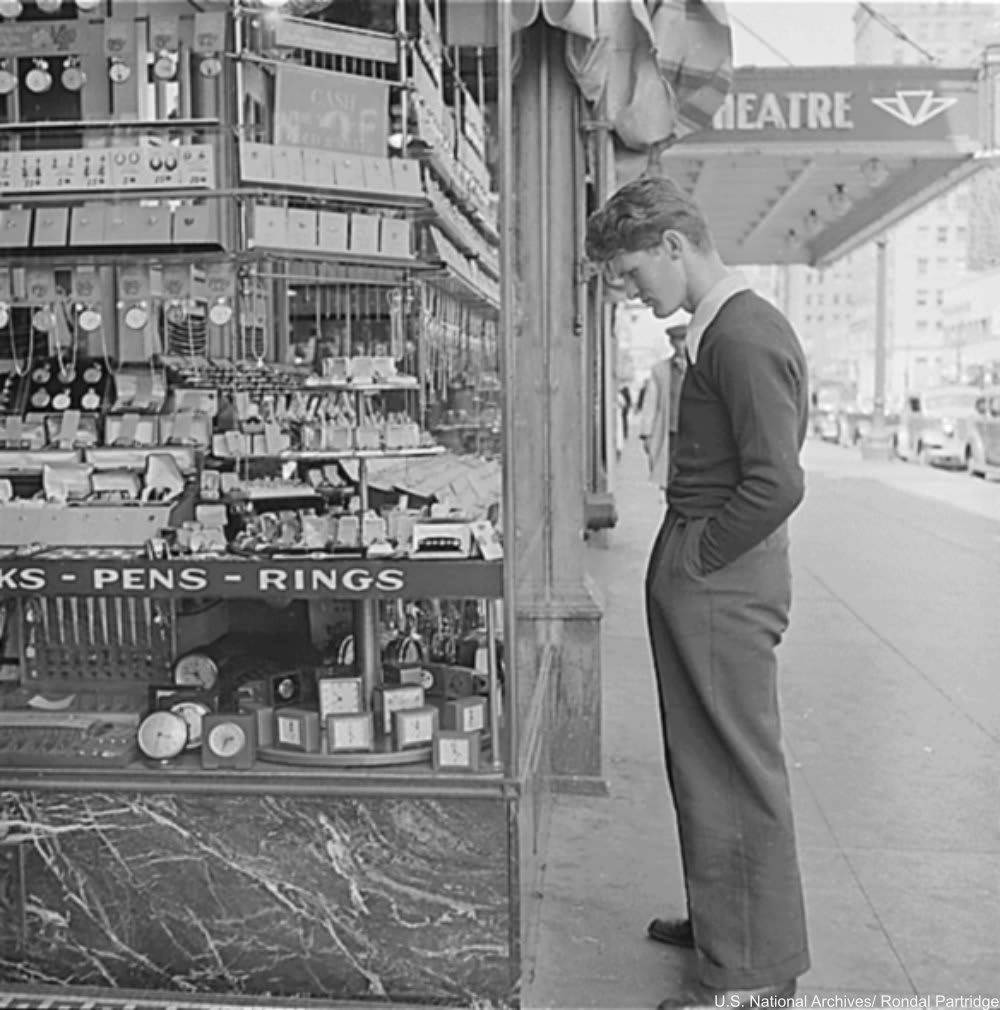 A young man in a sweater stands on a city sidewalk, looking down at items displayed in a shop window that sells clocks, pens, and rings. Theater signs and cars are visible in the background.