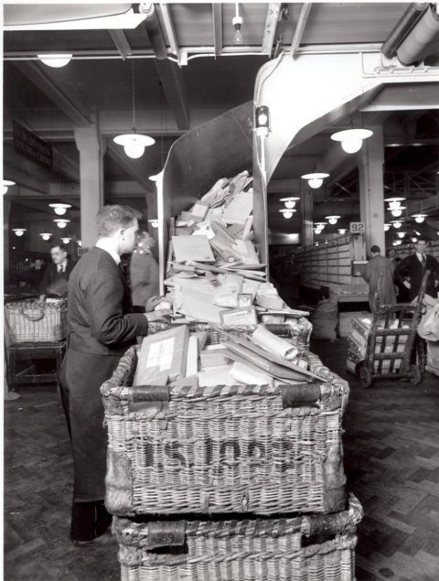 A man sorts through a large basket of mail inside a busy postal facility, surrounded by letters, packages, and postal carts under bright ceiling lights.