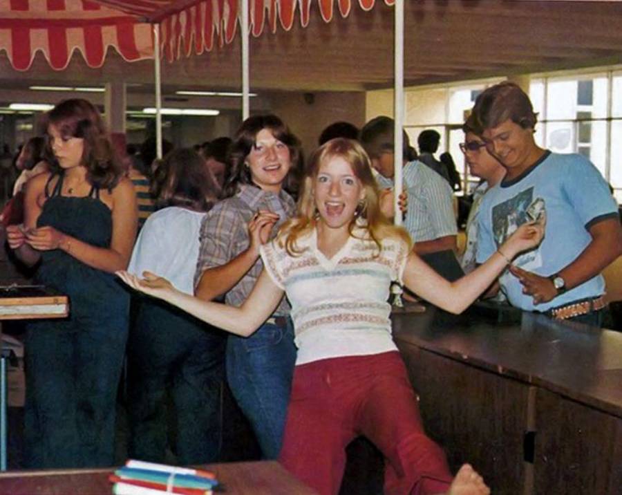 A young woman in a striped shirt and red pants poses playfully at the front of a line of smiling teens under a red-and-white canopy, inside a brightly lit room.