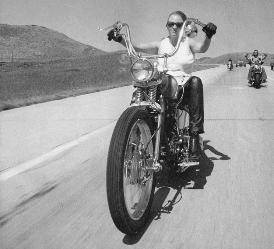 A person wearing sunglasses and gloves rides a large motorcycle on an open road, with others on motorcycles following behind.
