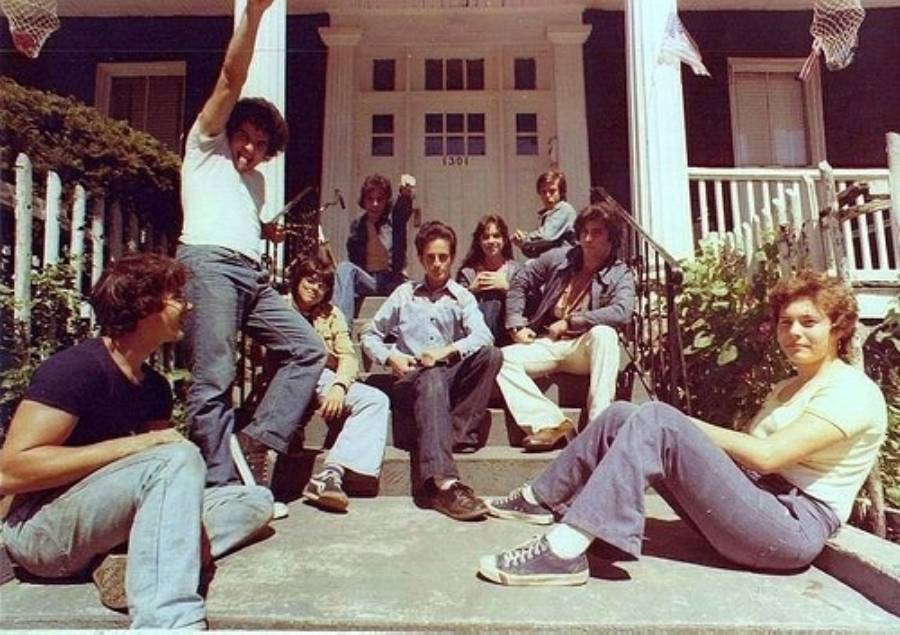 Nine young adults sit and pose casually on the front steps of a house with white columns and an address number "1301" above the door, looking relaxed and friendly on a sunny day.