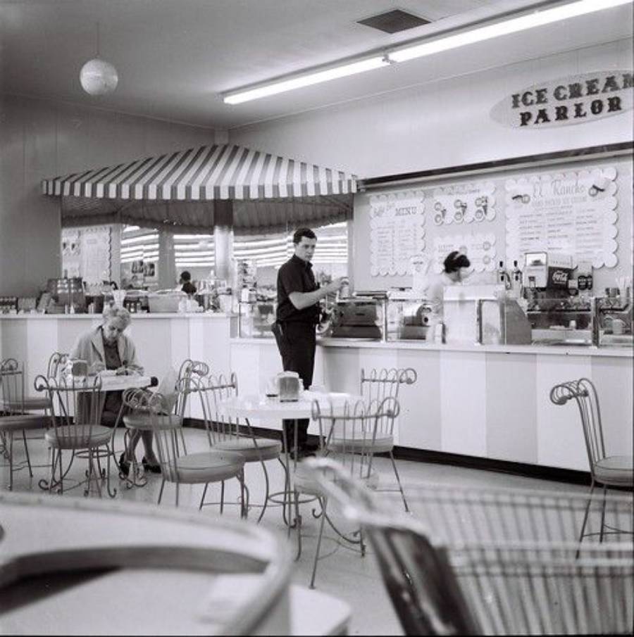 Black-and-white photo of a vintage ice cream parlor with a striped awning, menu boards, metal chairs, a man standing, a woman behind the counter, and a seated customer reading.