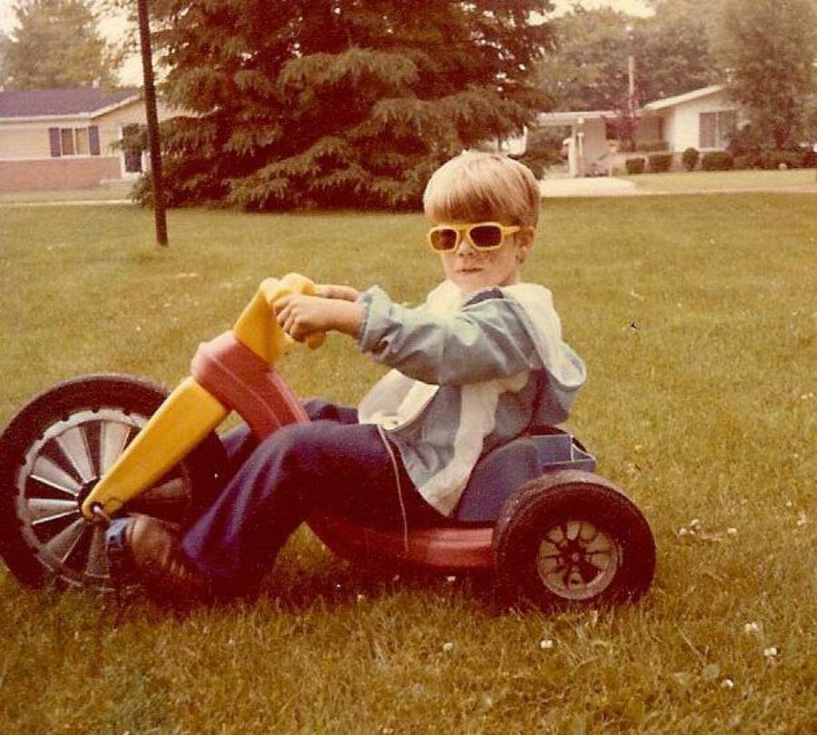 A child wearing yellow sunglasses rides a plastic tricycle on a grassy lawn in a suburban neighborhood with houses and trees in the background.