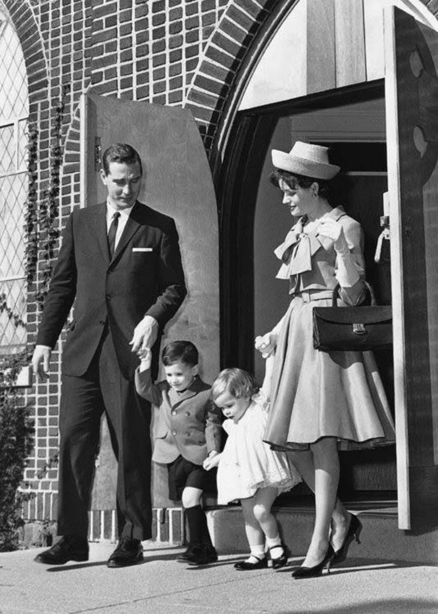 A man in a suit and a woman in a dress and hat stand outside a building with two young children, holding their hands as they step down from a doorway. The scene appears formal and vintage.