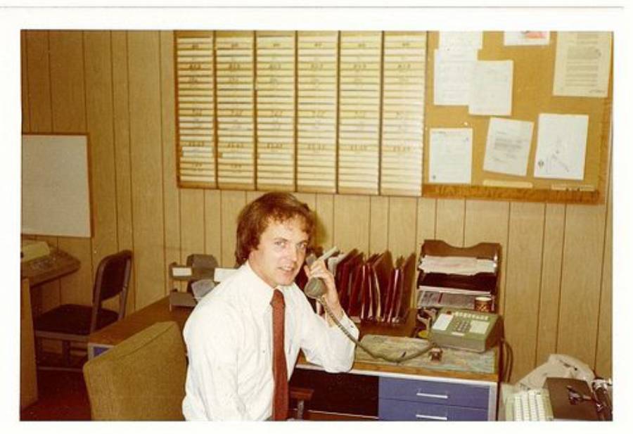 A man in a white shirt and tie sits at an office desk, speaking on a corded phone. Behind him are shelves with files and a bulletin board covered with papers and schedules. The office has wood-paneled walls.