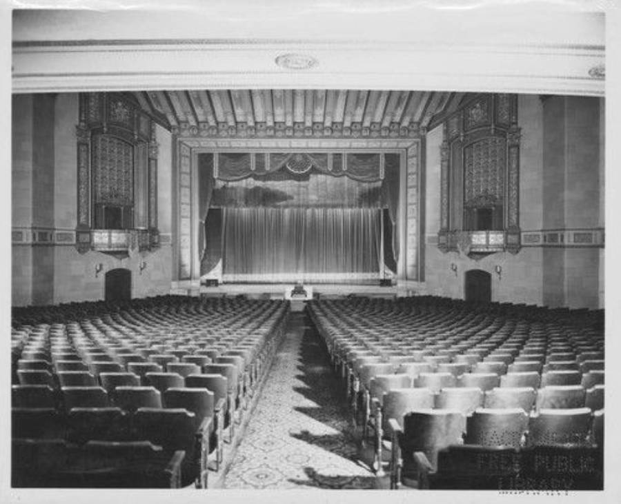 Black and white photo of an empty theater auditorium with rows of seats facing a stage with closed curtains. Ornate architectural details decorate the walls and ceiling.