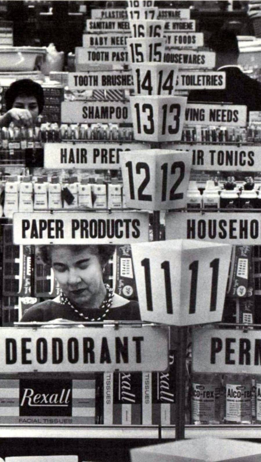 Black and white photo of two women shopping in a store aisle, surrounded by stacked aisle signs for products like paper products, deodorant, toiletries, hair products, and baby needs.