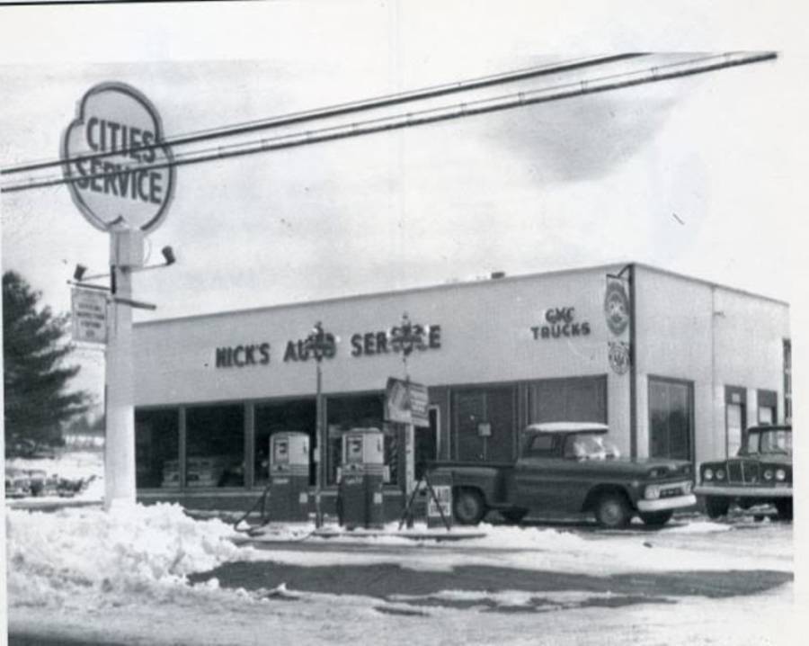 Black and white photo of a vintage gas station with “Nick’s Auto Service” on the building, two old gas pumps, a pickup truck, and snow on the ground. A large “Cities Service” sign is out front.