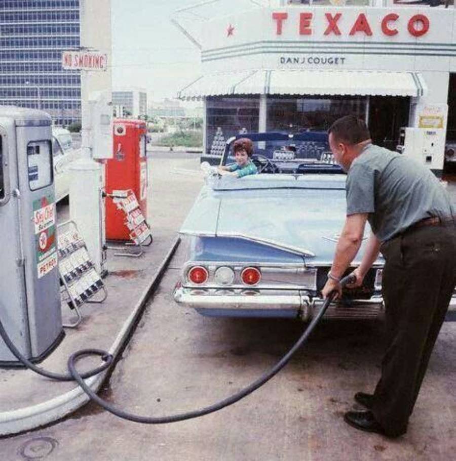 A man pumps gas into a vintage blue car at a Texaco station. A woman sits in the driver's seat, smiling. The scene has a retro feel, with old gas pumps and a diner in the background.