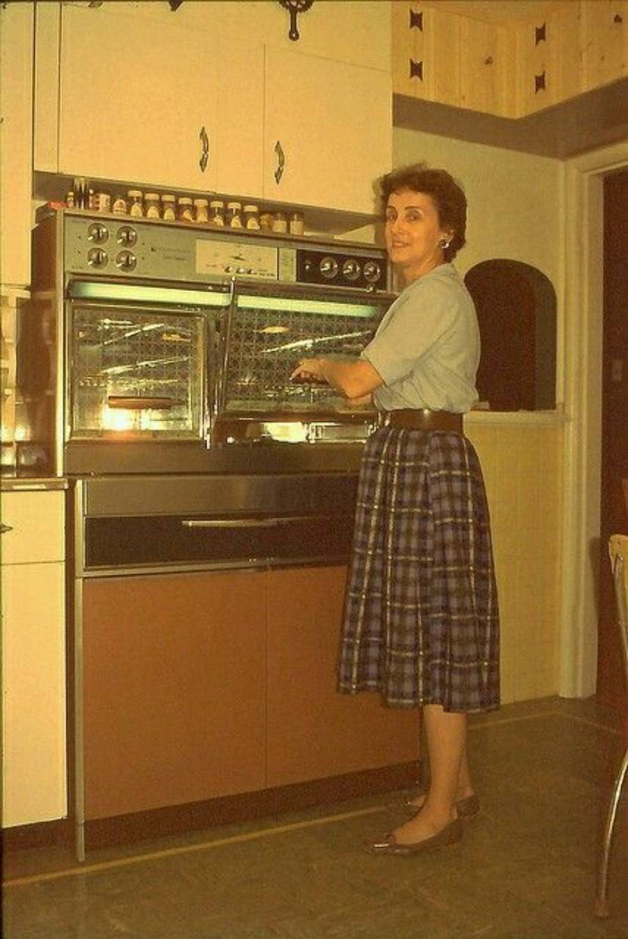 A woman in a plaid skirt and blouse stands in a retro kitchen, using a double oven. The room features warm, vintage colors and cabinetry.