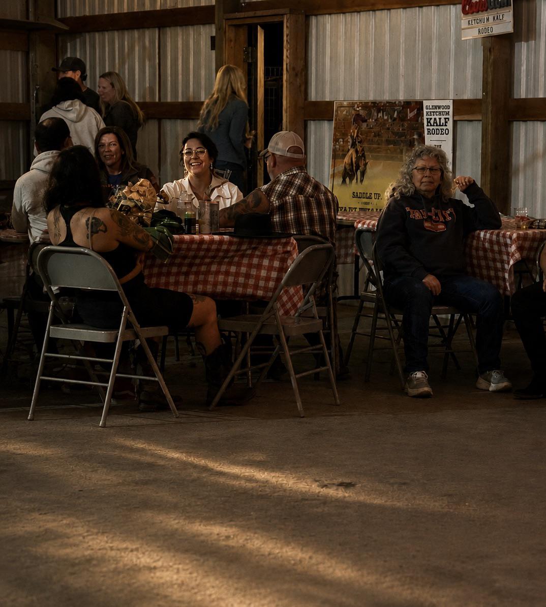 People sit at tables with checkered cloths inside a barn-like setting. Some talk and laugh, while one person sits alone. Sunlight streams onto the floor. Posters hang on a wood-and-metal wall in the background.