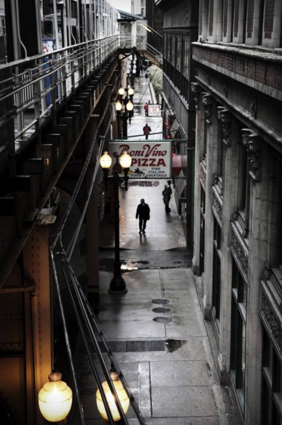 A narrow city alleyway beneath an elevated train track, with street lamps illuminating the wet pavement. A few people walk below, passing a sign for Roni Vino Pizza. Tall buildings line both sides of the alley.