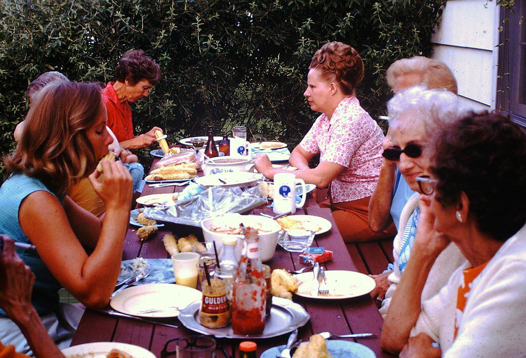 Several women sit around a picnic table outdoors, eating a meal that includes corn on the cob. The table is covered with plates, cups, condiments, and dishes, while greenery is visible in the background.