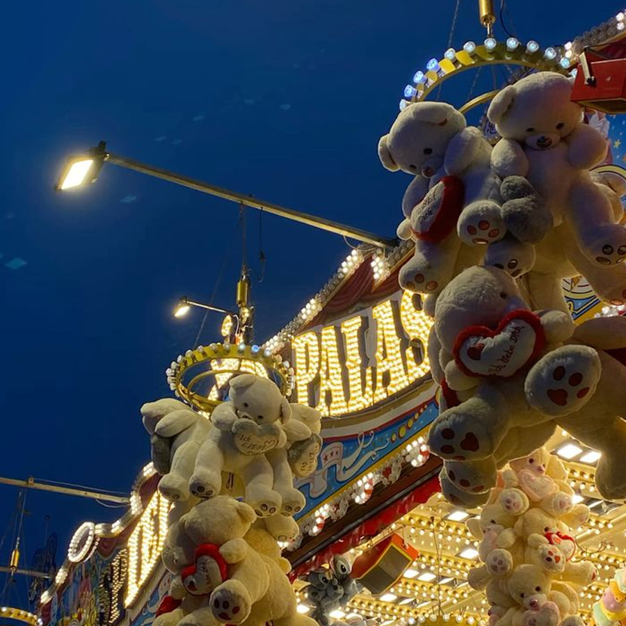 Stuffed teddy bears hang as prizes at a brightly lit carnival booth at night, with string lights and signage glowing against the dark blue sky.