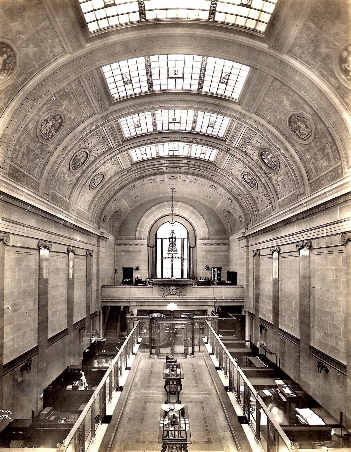 A grand historic bank interior with high vaulted ceilings, arched windows, ornate detailing, and rows of teller booths below. Natural light streams through large skylights above. Black-and-white photo.