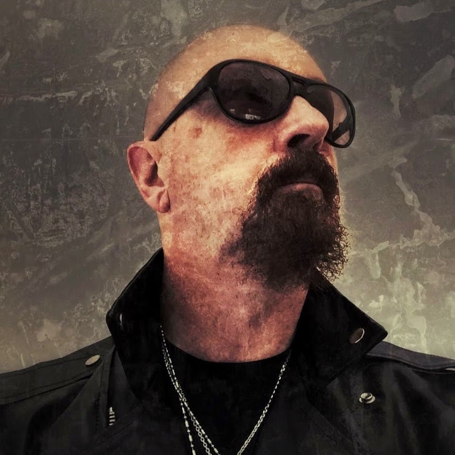 A person with a shaved head, sunglasses, and a goatee wears a black jacket with the collar up and chain necklaces, looking off to the side against a textured, grunge-style background.