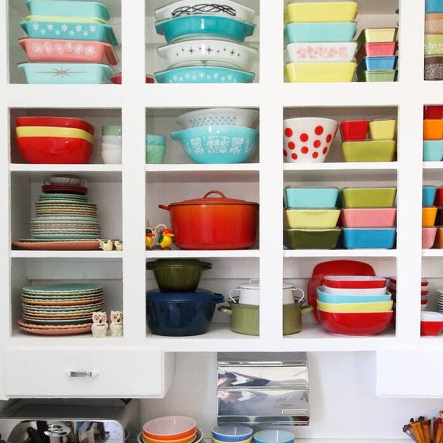 A white kitchen shelf filled with colorful vintage bowls, dishes, cookware, and mugs, neatly arranged in various cubbies. Some items have retro patterns and designs.