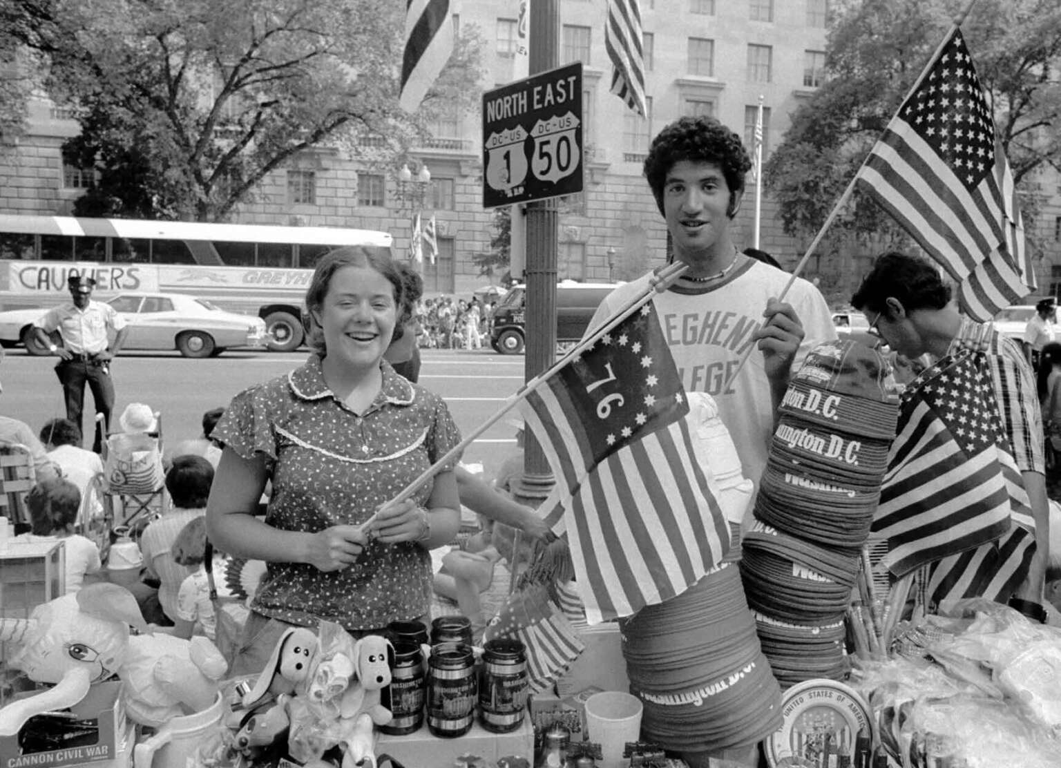Two people stand at a street market booth selling patriotic items, including U.S. flags and hats, during a parade. A bus, spectators, and trees are visible in the background. The mood is festive and cheerful.
