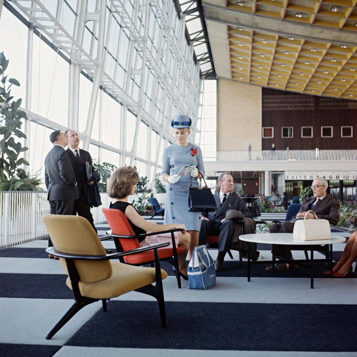 A vintage airport lounge with people in formal 1960s attire, including a flight attendant in a blue uniform and cap. Large windows, modern furniture, and a high yellow ceiling create a bright, spacious atmosphere.