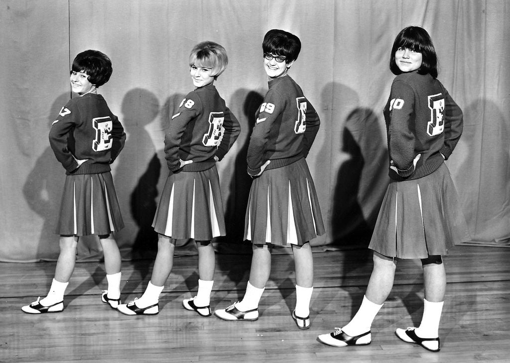 Four cheerleaders in matching sweaters, pleated skirts, and Mary Jane shoes pose in a line, slightly turned with hands on hips, in front of a curtain on a wooden floor. The image is in black and white.
