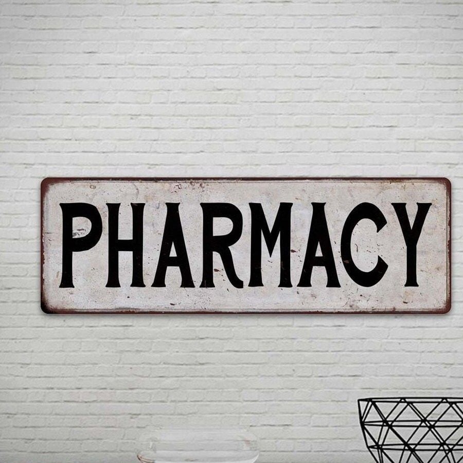 A rectangular sign with the word "PHARMACY" in bold black letters is mounted on a white brick wall. A metal wireframe object is partially visible in the bottom right corner.