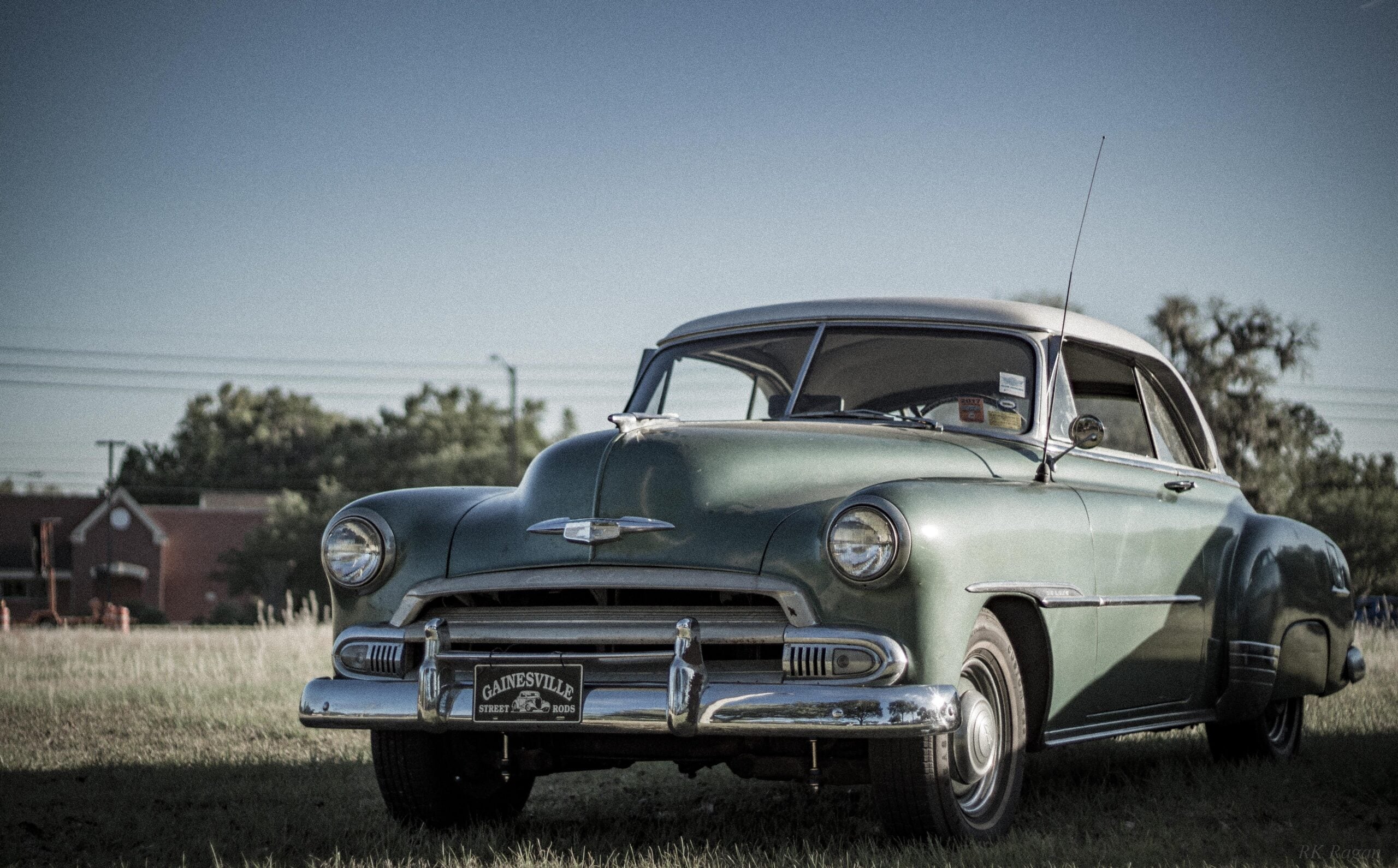 A vintage green car is parked on grass under a clear sky, with trees and a building in the background. The front license plate reads "Gainesville Street Rods.