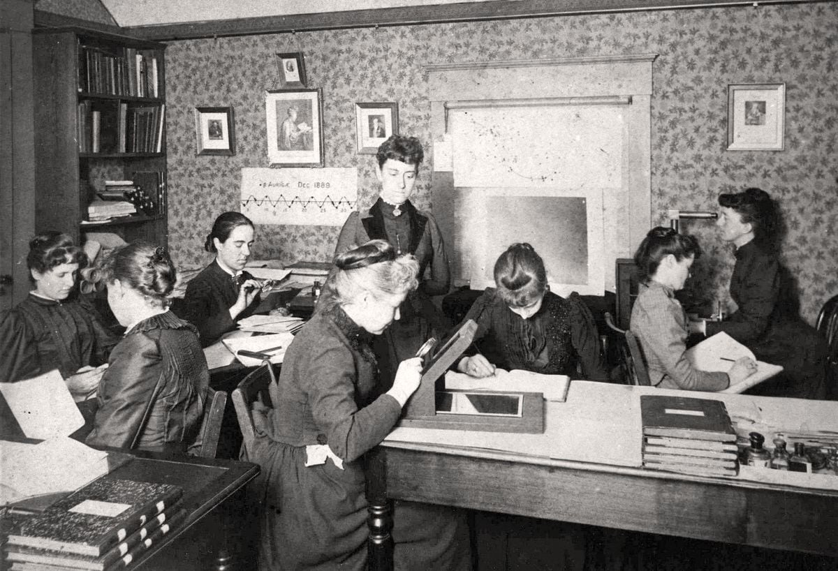 Eight women in Victorian-era clothing work at desks with papers, books, and drawing tools in a small, wallpapered office. Shelves and framed pictures decorate the walls. The scene is scholarly and focused.