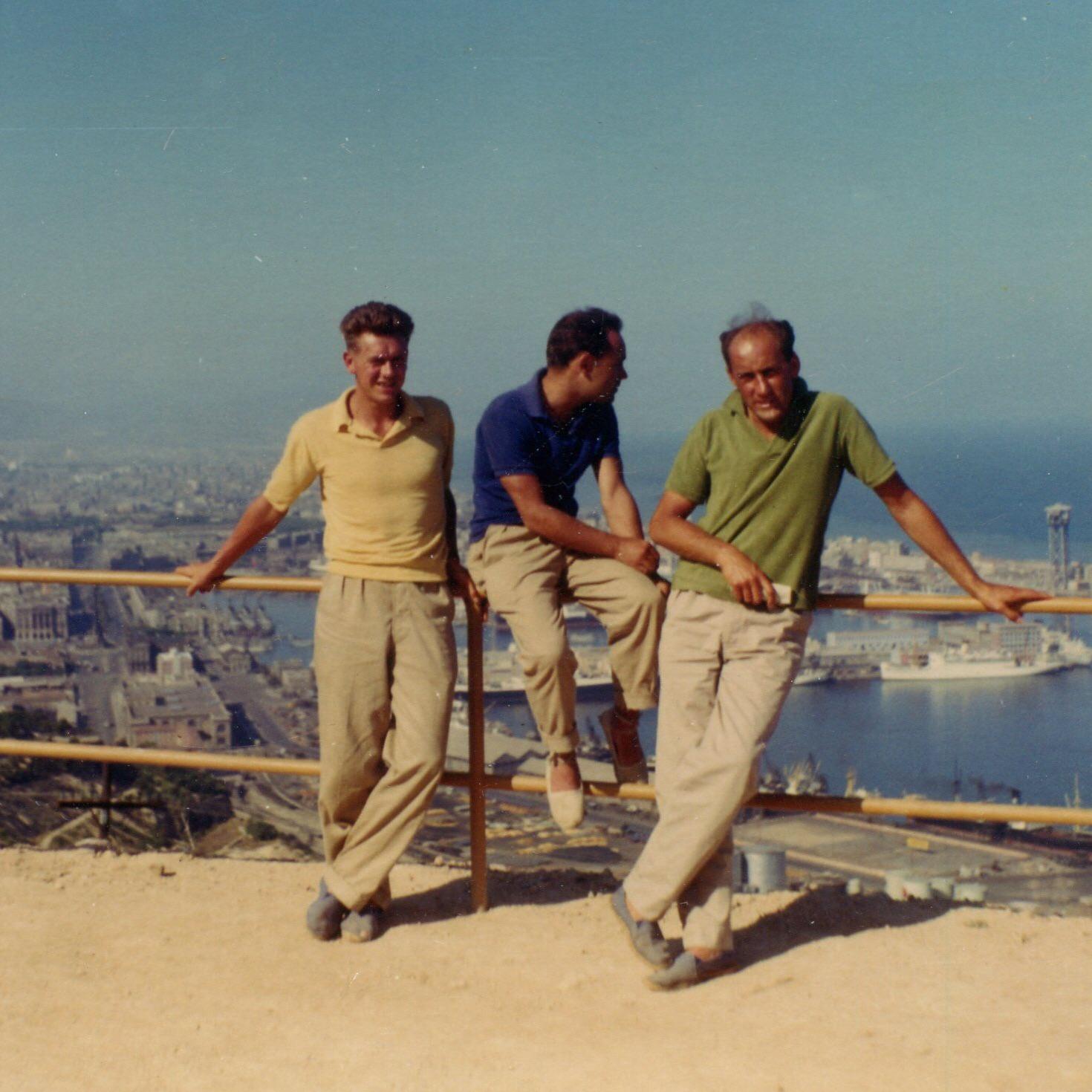 Three men in casual clothes pose by a wooden railing overlooking a coastal city and harbor on a sunny day. The background shows buildings, water, and a clear blue sky.
