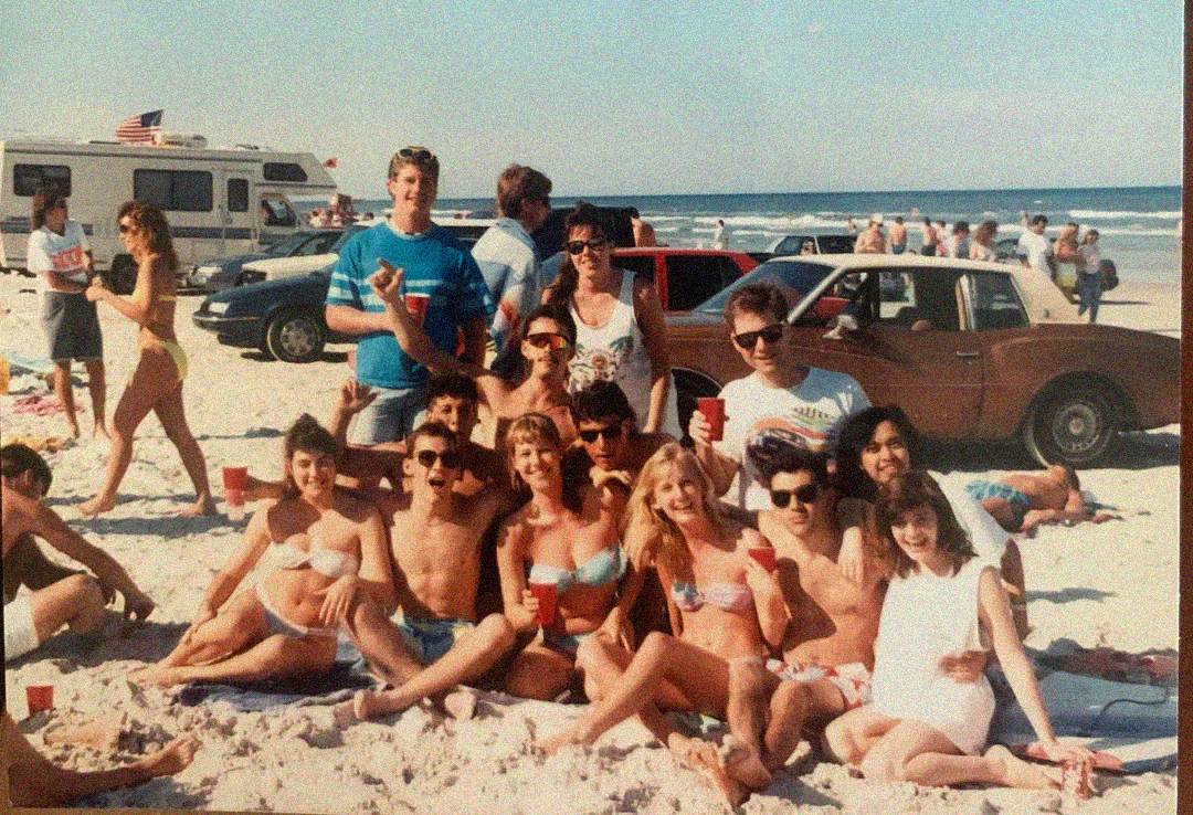 A group of young adults in swimsuits pose and smile for a photo on a sandy beach with red cups. Cars and an RV are parked in the background, and the ocean is visible under a clear sky.