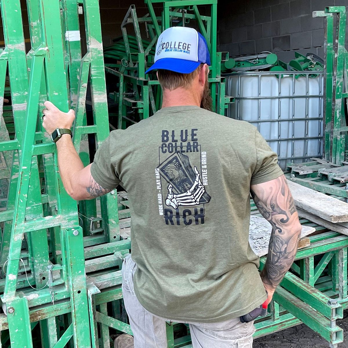 A man in a blue cap and olive T-shirt reading “BLUE COLLAR RICH” stands among green metal scaffolding, facing away from the camera with one hand resting on the structure.