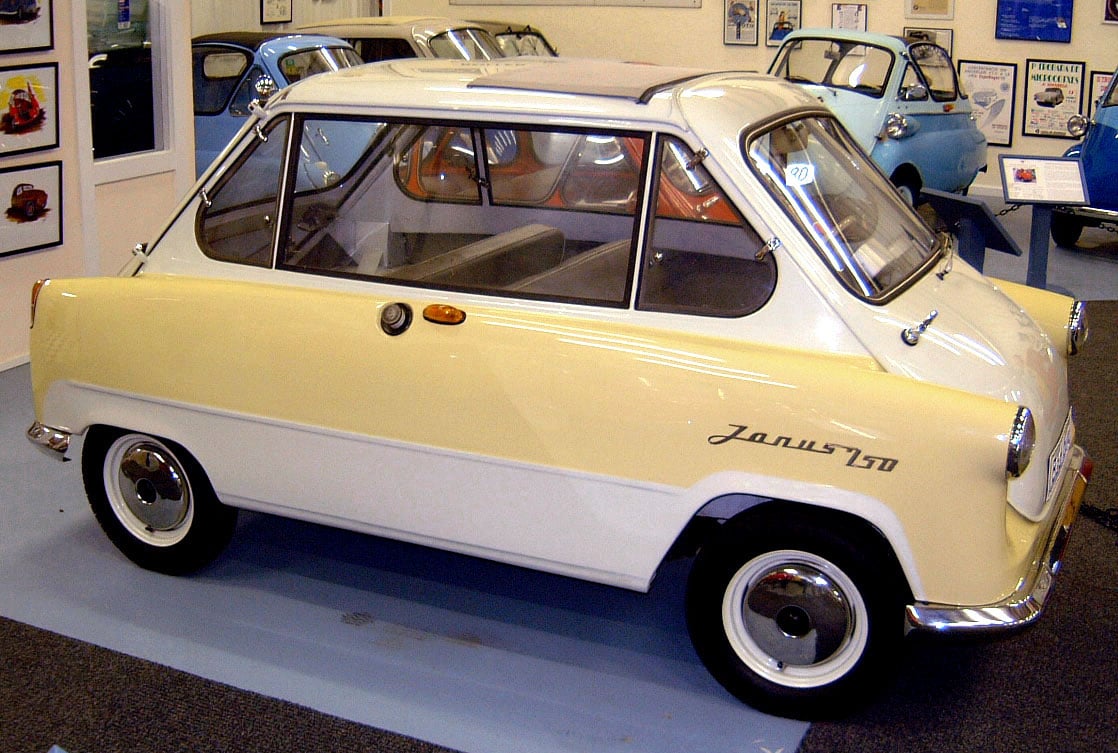 A vintage yellow and white Zündapp Janus 750 microcar is on display indoors, surrounded by other classic cars and automotive memorabilia.