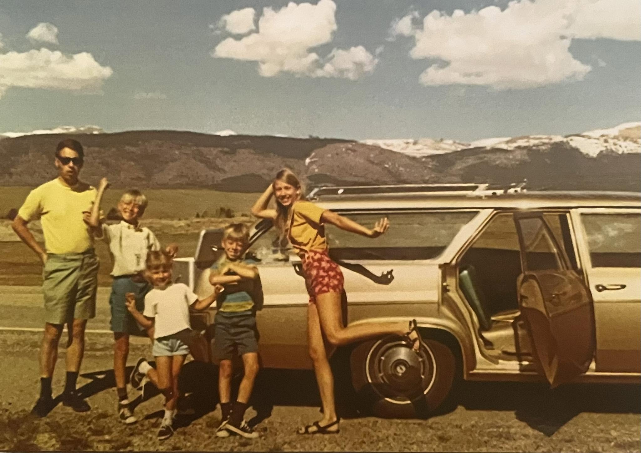 A family of five poses by a vintage station wagon on a scenic roadside, with mountains and clouds in the background. The car door is open and everyone is smiling, suggesting a joyful road trip.