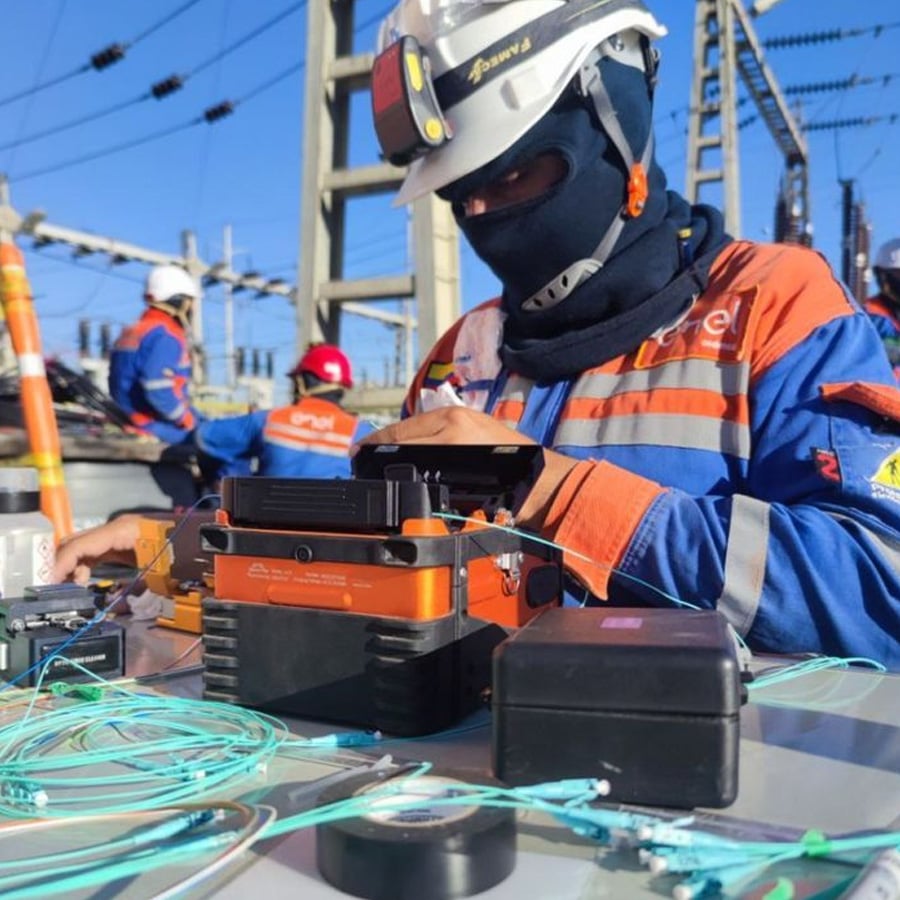 A worker in safety gear handles fiber optic cables and equipment at an outdoor electrical substation. Other workers are visible in the background among power lines and towers.