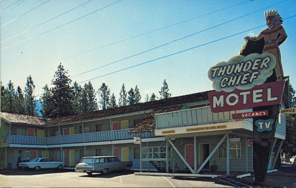 Vintage motel with a large sign reading "Thunder Chief Motel," featuring a figure in a headdress. Two classic cars are parked in front, with trees and blue sky in the background.