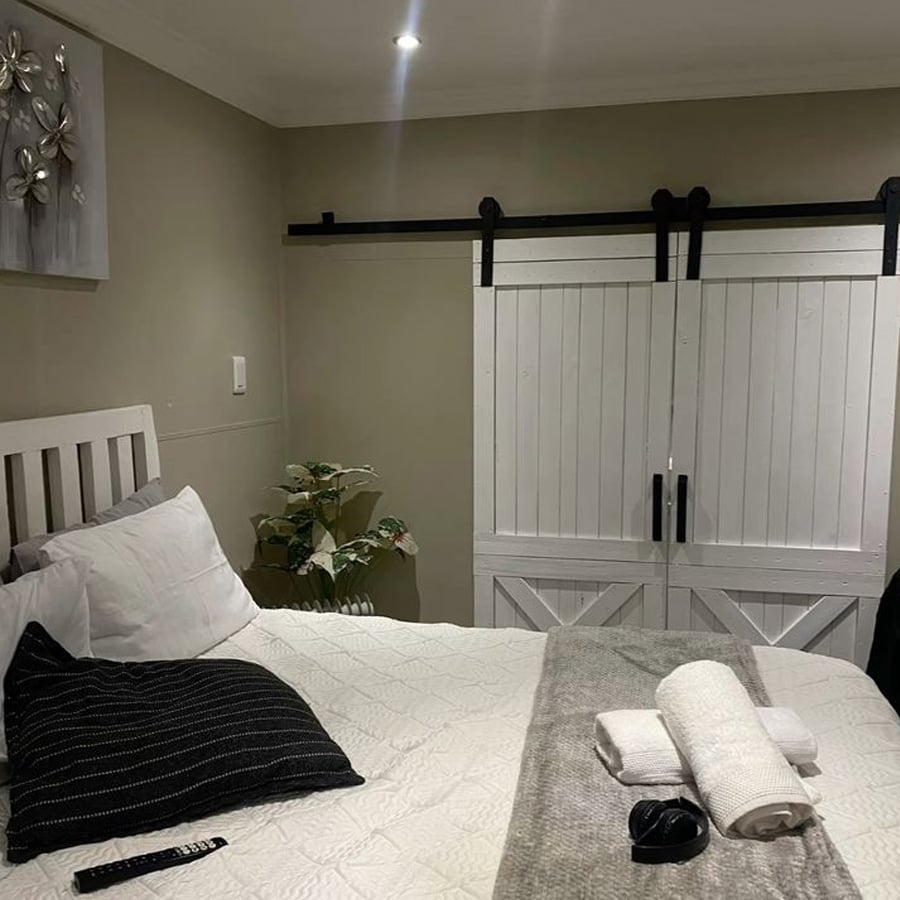 A cozy bedroom with a white bed, black and white pillows, folded towels, a remote control, a potted plant, and white sliding barn doors against a taupe wall. A floral wall art piece hangs above the bed.
