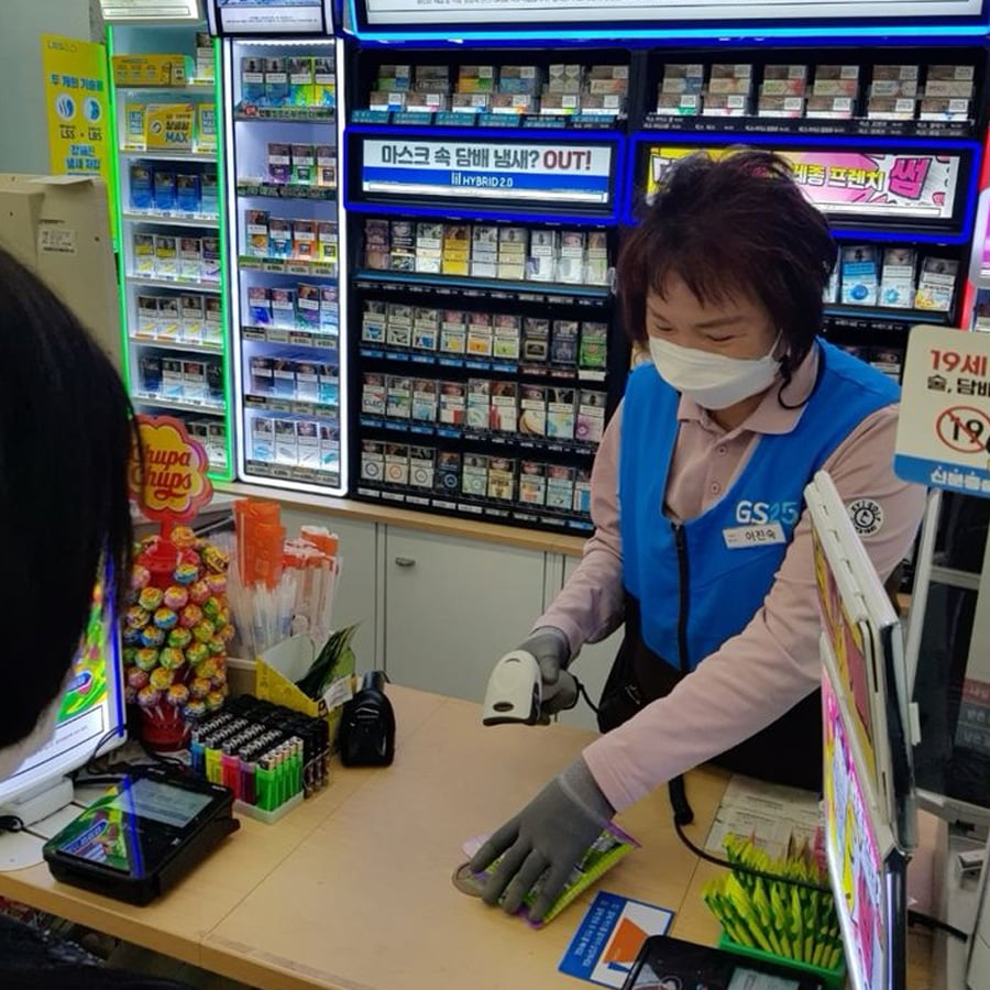 A cashier wearing a mask and gloves scans an item at a convenience store counter stocked with lollipops, candy, and tobacco products. Signs and cigarette packs are displayed behind her.
