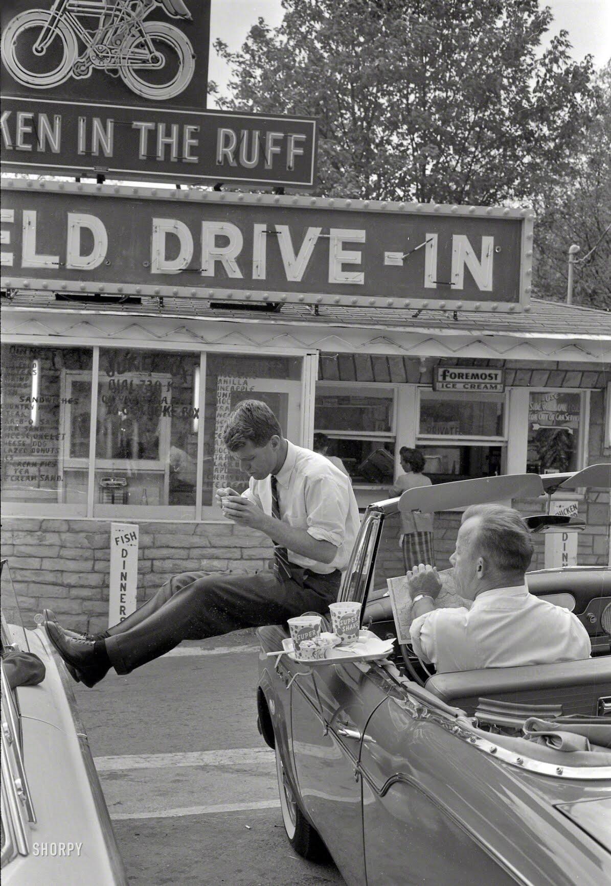 A young man in a suit sits on the open door of a convertible at a drive-in restaurant, eating food from a tray, while another man sits inside the car. The drive-in’s retro sign and menu are visible in the background.