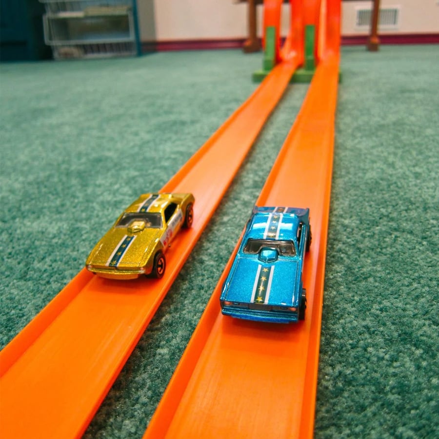 Two toy cars, one gold and one blue, race side by side on orange plastic tracks placed on a green carpet.