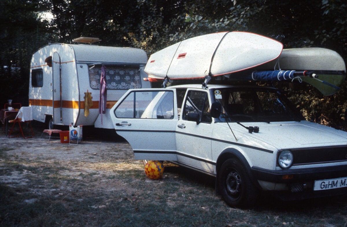 A white car with kayaks on the roof is parked by a white camper. The car’s door is open, and a colorful ball is on the grass nearby. Trees and camping items are in the background.