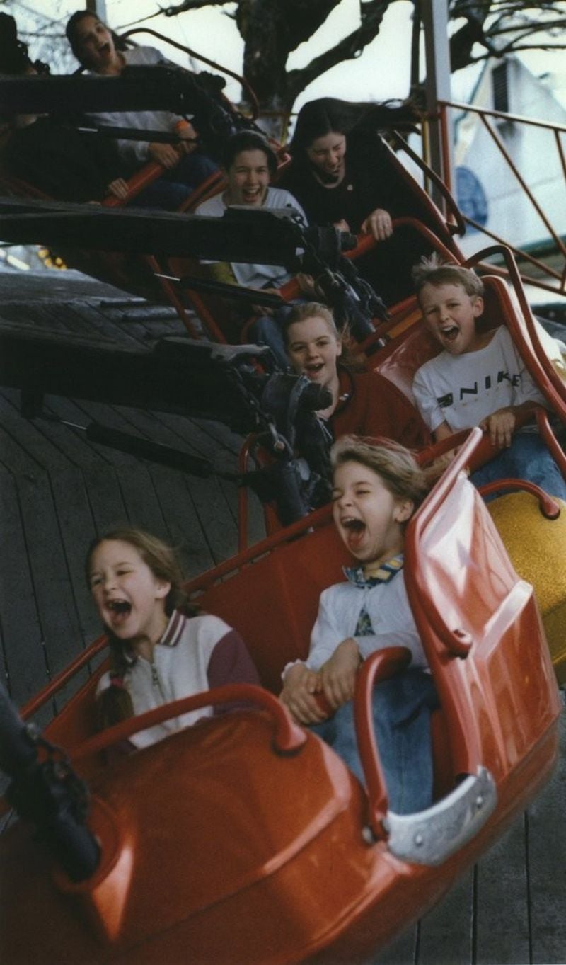 A group of children are laughing and screaming with excitement as they ride together on a spinning amusement park ride in bright red seats. The background shows part of the ride structure and trees.
