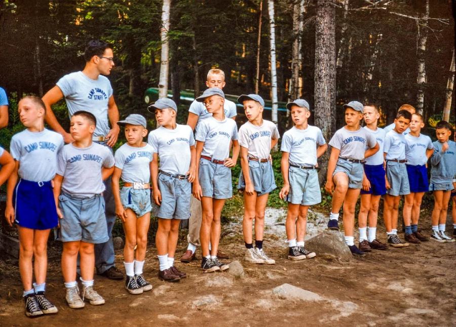 A group of boys in matching "Camp Sunshine" shirts and shorts stand in a line outdoors, supervised by two adults, with trees and dirt ground in the background.