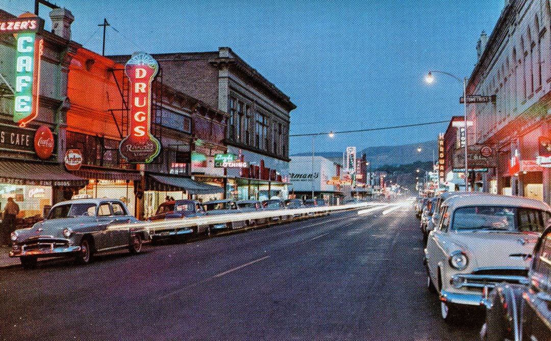 A vintage city street at dusk with neon signs for a cafe and a drugstore, classic cars parked along the curb, and bright headlights creating light trails in the middle of the road.