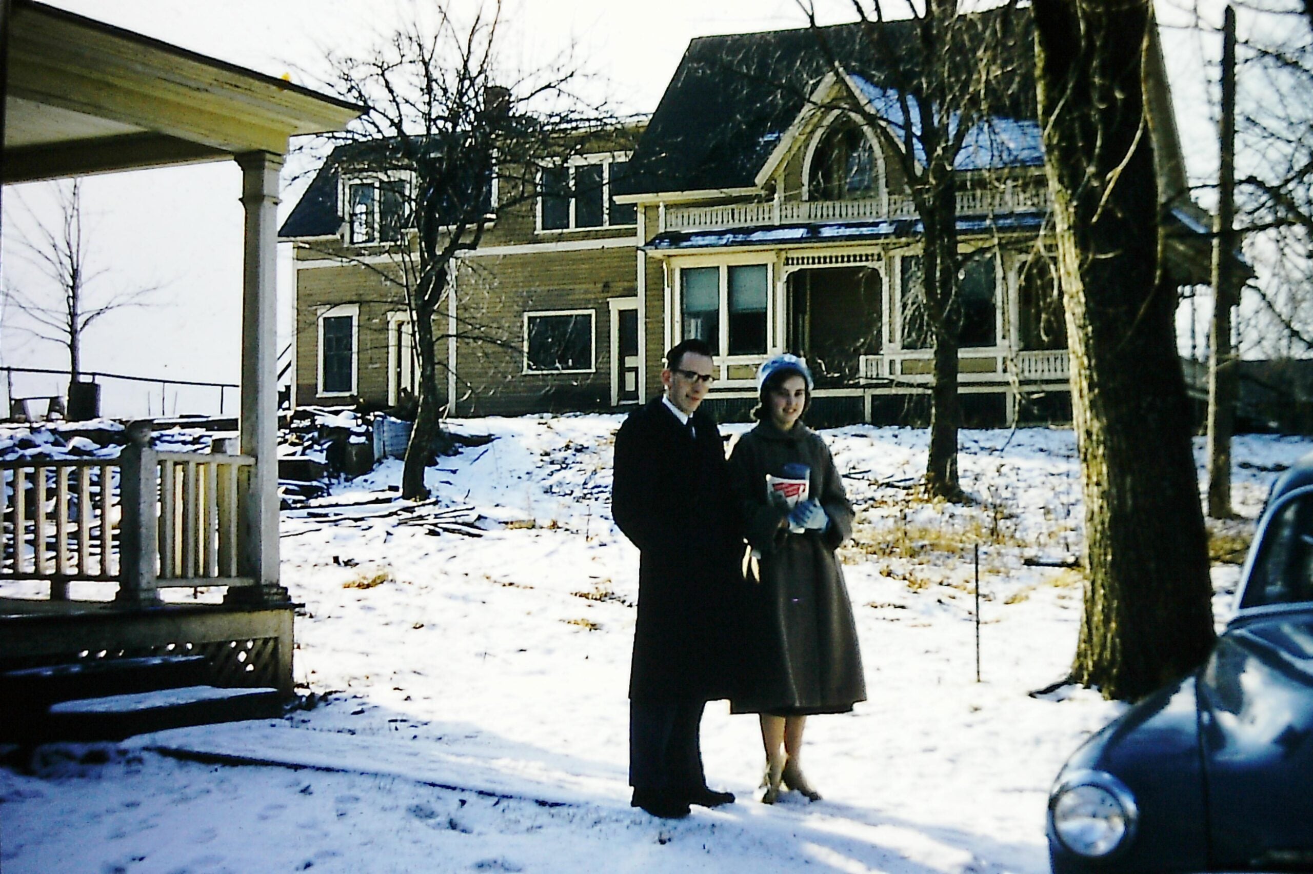 A man and woman in winter coats stand on a snowy yard in front of a large, old house with a porch. Bare trees surround them, and part of a car is visible on the right side of the image.