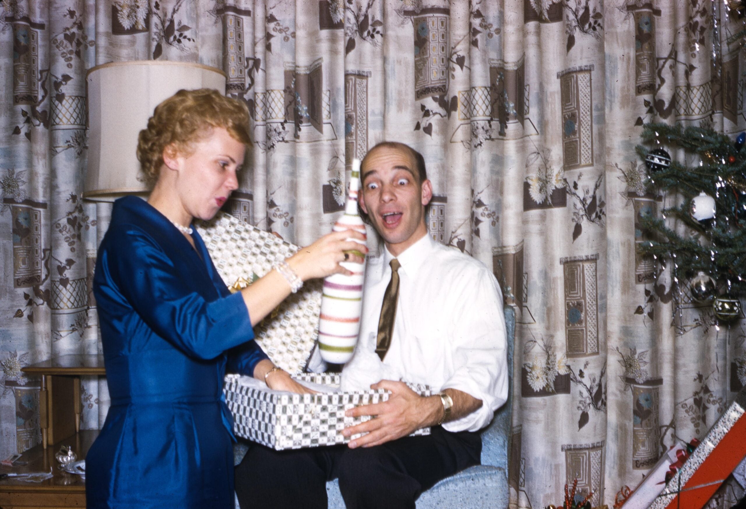 A woman in a blue dress hands a striped bottle to a surprised man with a gift box. A decorated Christmas tree and patterned curtains are in the background.