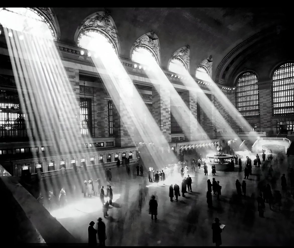 Black-and-white photo of a busy train station, with sunlight streaming through tall arched windows, casting dramatic beams of light onto people walking and standing on the main concourse.