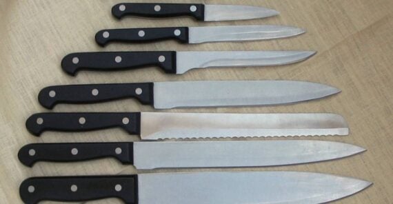 Eight kitchen knives of various sizes and types with black handles are arranged in a row on a beige fabric surface.