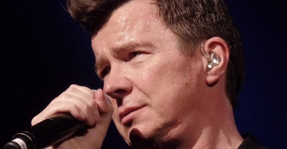 A man with short dark hair holds a microphone close to his mouth while singing on stage. He is wearing a black outfit and an in-ear monitor. The lighting highlights his face and expression.