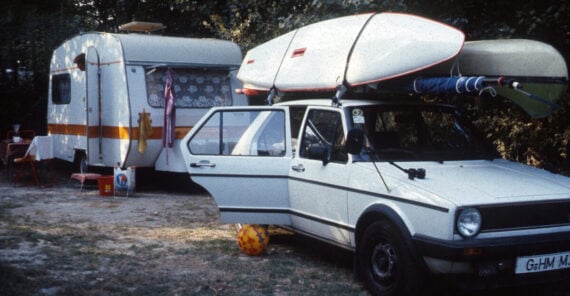 A white car with two kayaks on its roof is parked near a camper trailer in a grassy area. A soccer ball is near the car, and camping items are set up outside the trailer under the trees.